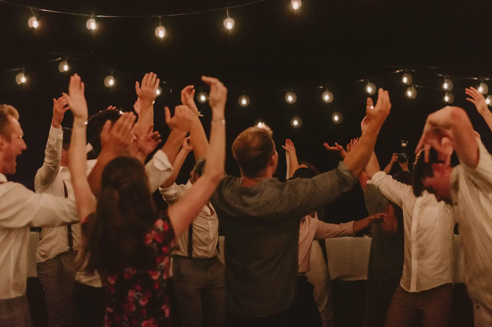 Convidados dançando de braços para cima sob um varal de luzes em uma festa à noite